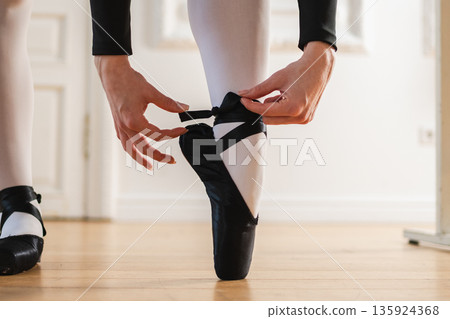 Ballerina puts on pointe shoes on leg in dance class. Classical ballet dancer woman in dance studio. Ballerina putting on tying classical pointe shoes for dance training. Ballet school concept Ballerina puts on pointe shoes on leg in dance class. Classical ballet dancer woman in dance studio. Ballerina putting on tying classical pointe shoes for dance training. Ballet school concept 135924368