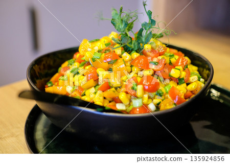 Fresh colorful corn salad with cherry tomatoes, bell pepper, green onions and herbs served in a black bowl on a rustic table. Healthy vegan summer side dish with vibrant vegetables and natural light. Fresh colorful corn salad with cherry tomatoes, bell pepper, green onions and herbs served in a black bowl on a rustic table. Healthy vegan summer side dish with vibrant vegetables and natural light. 135924856