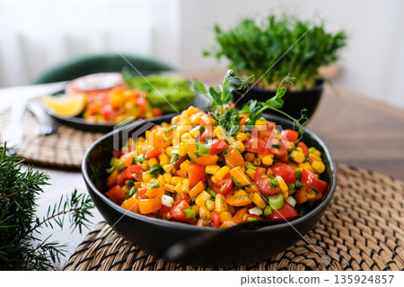 Fresh colorful corn salad with cherry tomatoes, bell pepper, green onions and herbs served in a black bowl on a rustic table. Healthy vegan summer side dish with vibrant vegetables and natural light. Fresh colorful corn salad with cherry tomatoes, bell pepper, green onions and herbs served in a black bowl on a rustic table. Healthy vegan summer side dish with vibrant vegetables and natural light. 135924857
