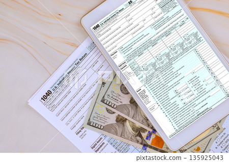 Digital tablet displays tax 1040 forms while US cash banknote is scattered on table with other tax documents. 135925043