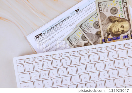 Tax 1040 return form on desk next to several one hundred dollar bills, bookkeeping work during financial tax preparations. 135925047