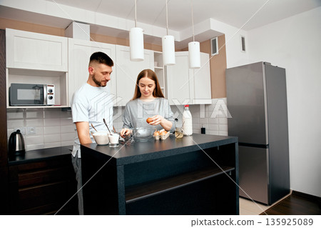 Couple prepares meal together in modern kitchen with bright lighting during morning hours Couple prepares meal together in modern kitchen with bright lighting during morning hours 135925089