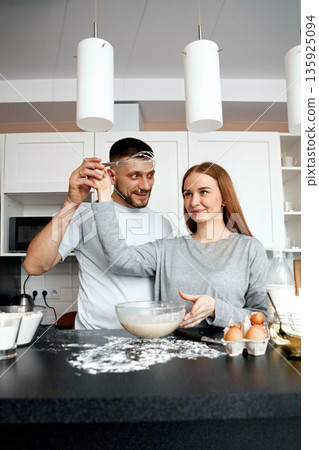 Couple interacts while preparing food in a kitchen with cooking supplies and ingredients scattered around during a casual cooking session 135925094