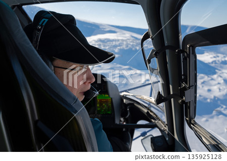 Pilot in a helicopter cockpit over snowy mountains, capturing fresh powder scenery 135925128