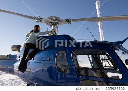 Woman climbs onto a blue helicopter in bright winter sun, preparing for a snow flight adventure Woman climbs onto a blue helicopter in bright winter sun, preparing for a snow flight adventure 135925132