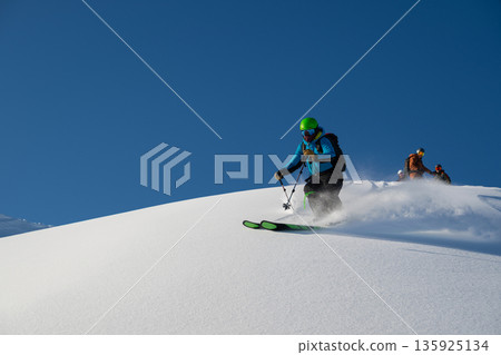 Off-piste freeride skiers navigate fresh powder on a blue-sky alpine slope. 135925134