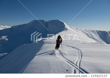 Off-piste freeride skiers traverse a snow-covered ridge under a clear blue sky in fresh powder on a high alpine peak 135925137