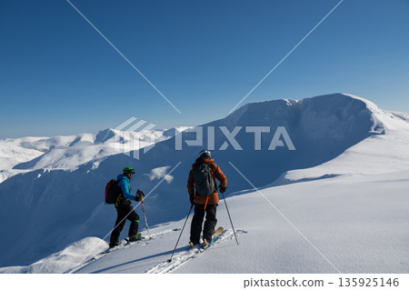 Two skiers traverse a snowy alpine ridge in clear blue sky during an off-piste adventure 135925146