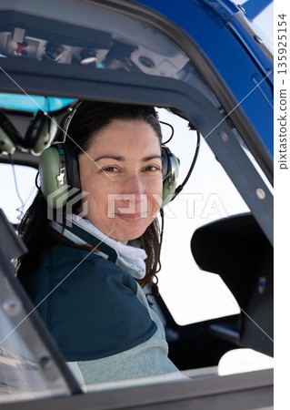 Female helicopter pilot wearing headset sits in cockpit, smiling in blue helicopter Female helicopter pilot wearing headset sits in cockpit, smiling in blue helicopter 135925154
