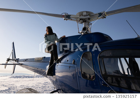 Woman sits on helicopter door beside cockpit in a blue chopper above a snowy landscape 135925157