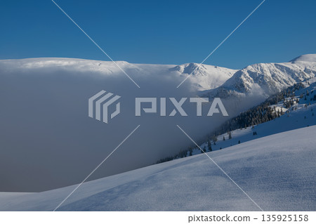 Snowy alpine mountains under blue sky with fog over pristine powder and expansive landscape Snowy alpine mountains under blue sky with fog over pristine powder and expansive landscape 135925158
