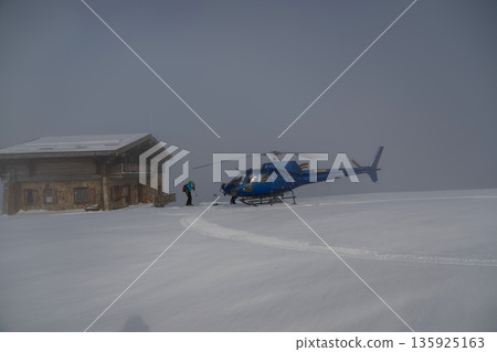 Blue helicopter on fresh powder beside a mountain lodge, ready for heliskiing adventure 135925163