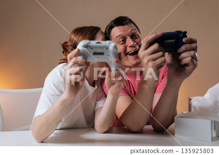 Happy excited couple bonding over online game, holding gaming controllers and having fun together while playing video game at home, sharing happy and joyful experience. 135925803