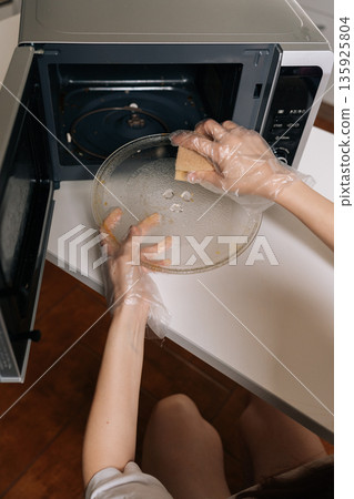 Top view of female hands in disposable gloves cleaning microwave oven interior with yellow sponge, maintaining kitchen hygiene standards and ensuring household cleanliness. 135925804