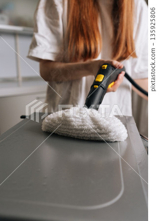 Vertical cropped shot of housewife cleaning kitchen microwave or appliance top with steam cleaner and white microfiber pad, producing visible steam to sanitize and deep clean surface. 135925806