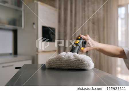 Hand using steam cleaner with fluffy white pad, removing dirt and germs from kitchen appliance surface, emphasizing eco-friendly and chemical-free cleaning concepts. Hand using steam cleaner with fluffy white pad, removing dirt and germs from kitchen appliance surface, emphasizing eco-friendly and chemical-free cleaning concepts. 135925811