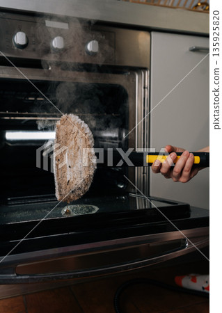 Housewife using steam cleaner with cleaning pad, generating steam and cleaning dirty stains inside kitchen oven, highlighting effective household chores and natural hygiene. 135925820