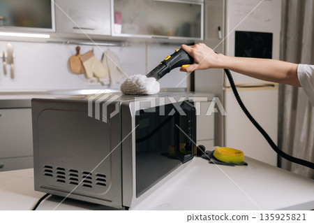 Closeup of cleaning service employee disinfecting microwave top with steam cleaner, removing grease and germs for spotless kitchen appliance. Concept of domestic hygiene and routine maintenance. 135925821