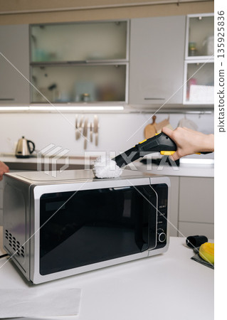 Closeup of unrecognizable housekeeper cleaning microwave top with steam cleaner, removing grease and germs for spotless kitchen appliance. Concept of domestic hygiene and routine maintenance. Closeup of unrecognizable housekeeper cleaning microwave top with steam cleaner, removing grease and germs for spotless kitchen appliance. Concept of domestic hygiene and routine maintenance. 135925836
