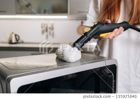 Close-up cropped shot of housewife cleaning microwave top with steam cleaner, removing grease and germs for spotless kitchen appliance. Concept of domestic hygiene and routine maintenance. Close-up cropped shot of housewife cleaning microwave top with steam cleaner, removing grease and germs for spotless kitchen appliance. Concept of domestic hygiene and routine maintenance. 135925840