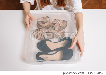 Top view of young adult woman organizing footwear, placing different pairs of shoes into clear plastic container, practicing home decluttering and efficient storage solution for tidiness. Top view of young adult woman organizing footwear, placing different pairs of shoes into clear plastic container, practicing home decluttering and efficient storage solution for tidiness. 135925852