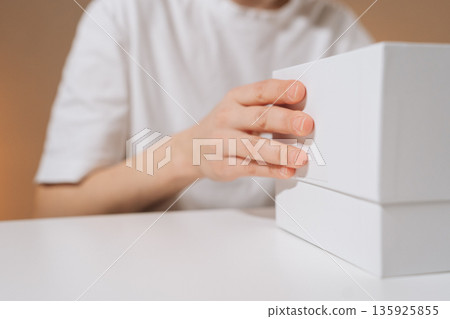 Person hand lifting lid off plain white box on minimalist white table, unboxing new electronic device or gift, revealing modern wireless game controller. Concept of modern gaming tech, leisure. Person hand lifting lid off plain white box on minimalist white table, unboxing new electronic device or gift, revealing modern wireless game controller. Concept of modern gaming tech, leisure. 135925855