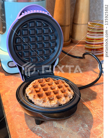A freshly baked waffle sits in an open electric waffle maker on a kitchen countertop, crisp golden texture contrasts with matte black plates A freshly baked waffle sits in an open electric waffle maker on a kitchen countertop, crisp golden texture contrasts with matte black plates 135925882