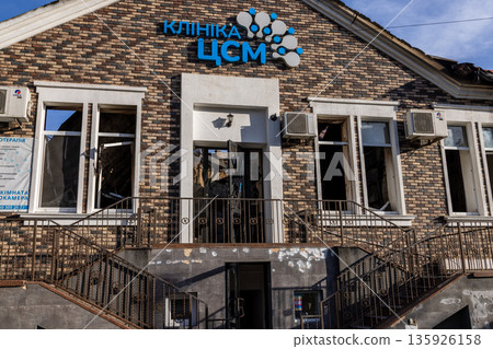 Brick clinic with Cyrillic signage shows broken windows and exterior damage; air conditioners and plants remain intact under clear blue sky. Brick clinic with Cyrillic signage shows broken windows and exterior damage; air conditioners and plants remain intact under clear blue sky. 135926158