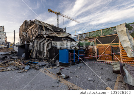 A heavily damaged building shows signs of fire and explosion collapsed roof charred walls scattered debris and a blue industrial unit wrapped in caution tape beneath a looming construction crane. A heavily damaged building shows signs of fire and explosion collapsed roof charred walls scattered debris and a blue industrial unit wrapped in caution tape beneath a looming construction crane. 135926163