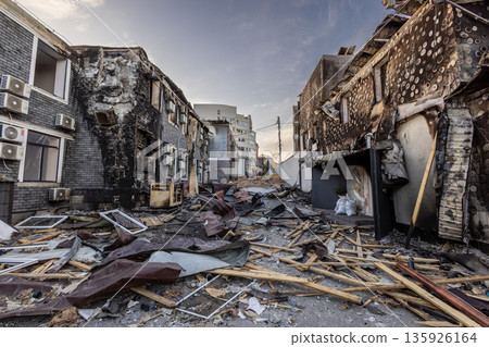 Two fire damaged buildings flank a debris strewn street charred walls broken windows and remnants of insulation mark the aftermath of destruction. 135926164