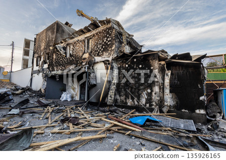 Severely damaged building with charred walls, collapsed sections, and scattered debris including a broken solar panel circular burn patterns mark the facade. Severely damaged building with charred walls, collapsed sections, and scattered debris including a broken solar panel circular burn patterns mark the facade. 135926165