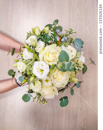 White floral bouquet held by hands, featuring roses, lisianthus, and greenery against a light wooden background 135926184