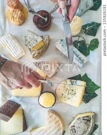 Assorted French cheese platter with various types of cheese, knife, and jam, hands preparing to serve Assorted French cheese platter with various types of cheese, knife, and jam, hands preparing to serve 135926191