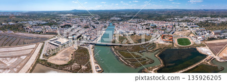 Aerial panorama view of Tavira highlighting the Gilao river, surrounding buildings, and open land in the Algarve region of Portugal under a clear blue sky 135926205