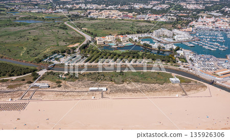 The marina at Vilamoura in Algarve Portugal with waterfront properties, green areas, and sandy beaches visible in the distance. Aerial 135926306