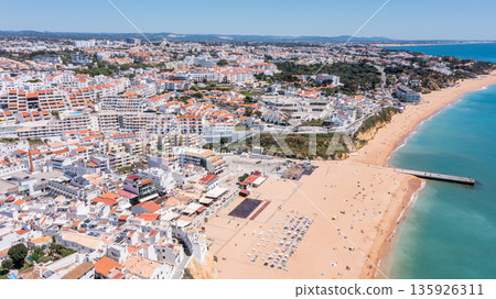 People enjoy the sandy shores and marina in Albufeira, Portugal. Beachgoers relax under the sun while boats dot the water. The town is vibrant and lively 135926311