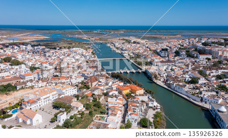 A drone view captures Tavira in Algarve with rivers, Roman bridges, and various structures. The coastline and landscapes are visible in the background 135926326