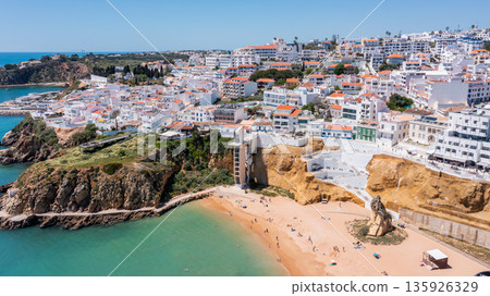 Tourists visit Albufeira in Portugal, Sunlit aerial view of coastal Albufeira in Portugal with whitewashed buildings on hills, orange rooftops, and beachgoers on golden sand near turquoise water. Tourists visit Albufeira in Portugal, Sunlit aerial view of coastal Albufeira in Portugal with whitewashed buildings on hills, orange rooftops, and beachgoers on golden sand near turquoise water. 135926329