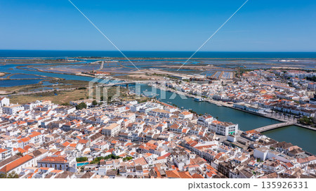 Coastal town Tavira Portugal with terracotta rooftops along winding river opening into wetlands and blue sea under sunny clear sky Concept of historic Mediterranean architecture and relaxed life Coastal town Tavira Portugal with terracotta rooftops along winding river opening into wetlands and blue sea under sunny clear sky Concept of historic Mediterranean architecture and relaxed life 135926331