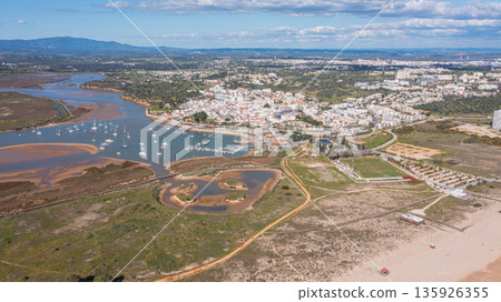 Aerial view of picturesque coastal town with boats in harbor, lush greenery, and sandy beach, showcasing vibrant tourism in Algarve region. Alvor Portugal Algarve Aerial view of picturesque coastal town with boats in harbor, lush greenery, and sandy beach, showcasing vibrant tourism in Algarve region. Alvor Portugal Algarve 135926355