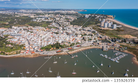 Aerial view of coastal town with vibrant buildings, boats in harbor, and scenic landscape showcasing tourism in a picturesque destination. Alvor Portugal Algarve Aerial view of coastal town with vibrant buildings, boats in harbor, and scenic landscape showcasing tourism in a picturesque destination. Alvor Portugal Algarve 135926359