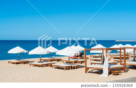 People lounge under white umbrellas on the sand at the beach. The sun shines brightly in the sky, and calm waves lap at the shore 135926368