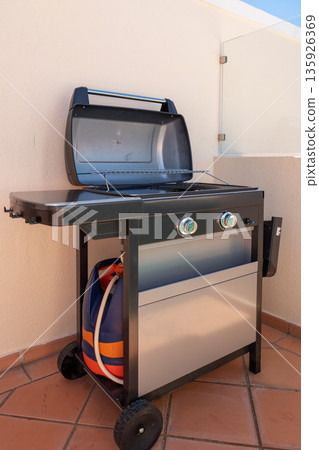 A grill sits on a patio terrace with its lid open, revealing the cooking surface and gas connections. It is positioned for outdoor cooking during the day 135926369