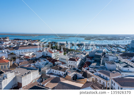 Tourists explore Faro's Old Town while boats float in the blue waters of Ria Formosa. The clear sky highlights the beauty of the Algarve region 135926370