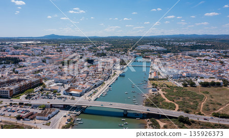 Drone captures Tavira in Algarve with Gilao river flowing through the town. Roads and boats are visible along the water, showcasing local life and scenery Drone captures Tavira in Algarve with Gilao river flowing through the town. Roads and boats are visible along the water, showcasing local life and scenery 135926373