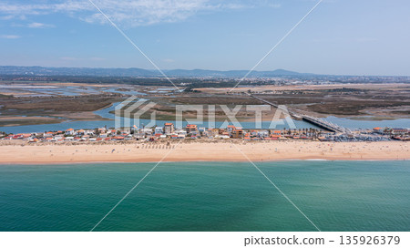 The sandy beach of Praia de Faro along with the wetlands of Ria Formosa. The coastline features various buildings and a long pier The sandy beach of Praia de Faro along with the wetlands of Ria Formosa. The coastline features various buildings and a long pier 135926379