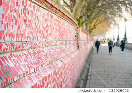 Visitors walk along the National Covid Memorial Wall in London. The wall is covered with red hearts, symbolizing loss and remembrance during the pandemic. Many people stop to reflect. Visitors walk along the National Covid Memorial Wall in London. The wall is covered with red hearts, symbolizing loss and remembrance during the pandemic. Many people stop to reflect. 135926602