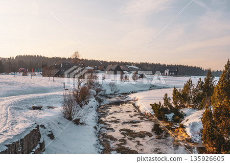 The winter sun rises over Jizerka village, illuminating cozy cottages and a gentle stream. Snow blankets the landscape, creating a serene and peaceful winter scene. The winter sun rises over Jizerka village, illuminating cozy cottages and a gentle stream. Snow blankets the landscape, creating a serene and peaceful winter scene. 135926605