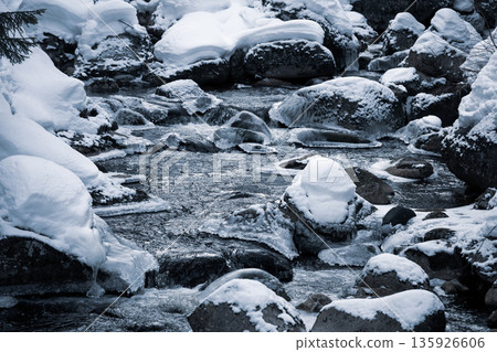 A tranquil winter scene features a cold river flowing through rocks covered in snow. The icy landscape captures the serene beauty of winter in the Czech Republic. A tranquil winter scene features a cold river flowing through rocks covered in snow. The icy landscape captures the serene beauty of winter in the Czech Republic. 135926606