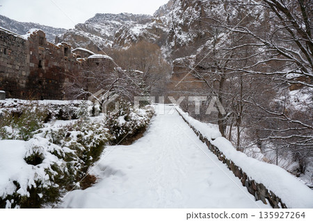 Snow-covered path leads toward stone walls of Geghard Monastery, framed by bare trees and steep rocky cliffs in muted winter light 135927264